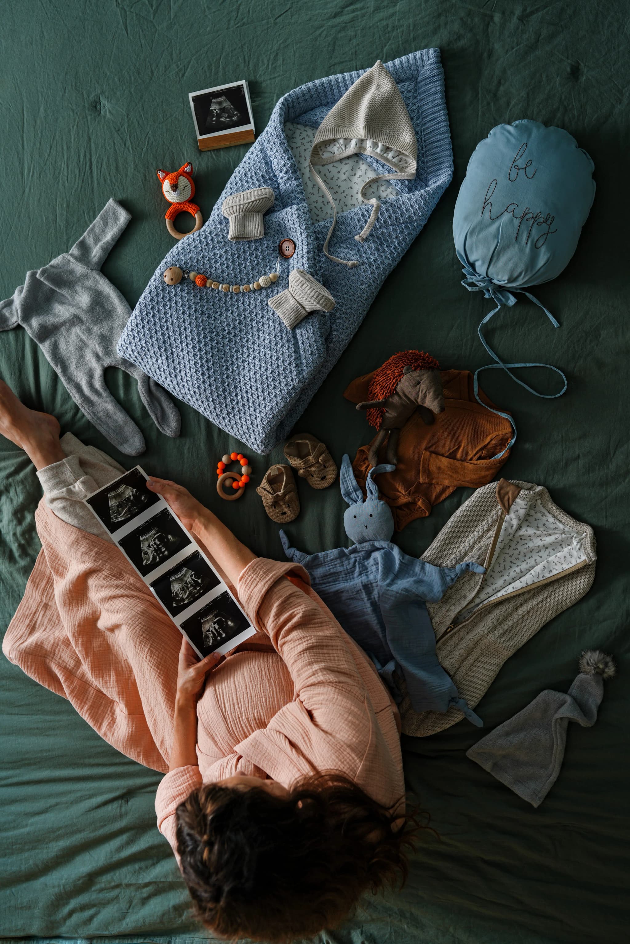 Future maman tenant des échographies, entourée de vêtements et accessoires pour bébé sur un lit.